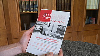 Zwei Hände halten Druckausgabe von Allemagne d’aujourd’hui, auf der man das Thema sieht. Im Hintergrund stehen Bücherregale.